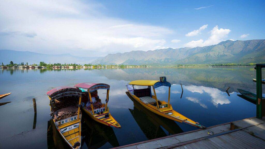 A serene lake with clear water reflects the blue sky and surrounding mountains.The distant shore is lined with trees , perfect for a Kashmir tour package.