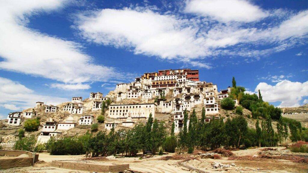 A white-washed monastery on a hillside under a blue sky, surrounded by lush trees and rocky terrain—typical of Ladakh trip packages.